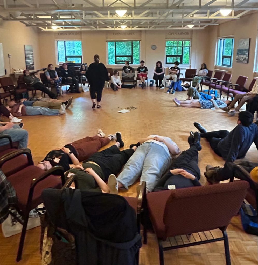 A circle of chairs in a room. Some people are seated on the chairs some are lying down on the floor. Maureen is standing in the middle of the circle.